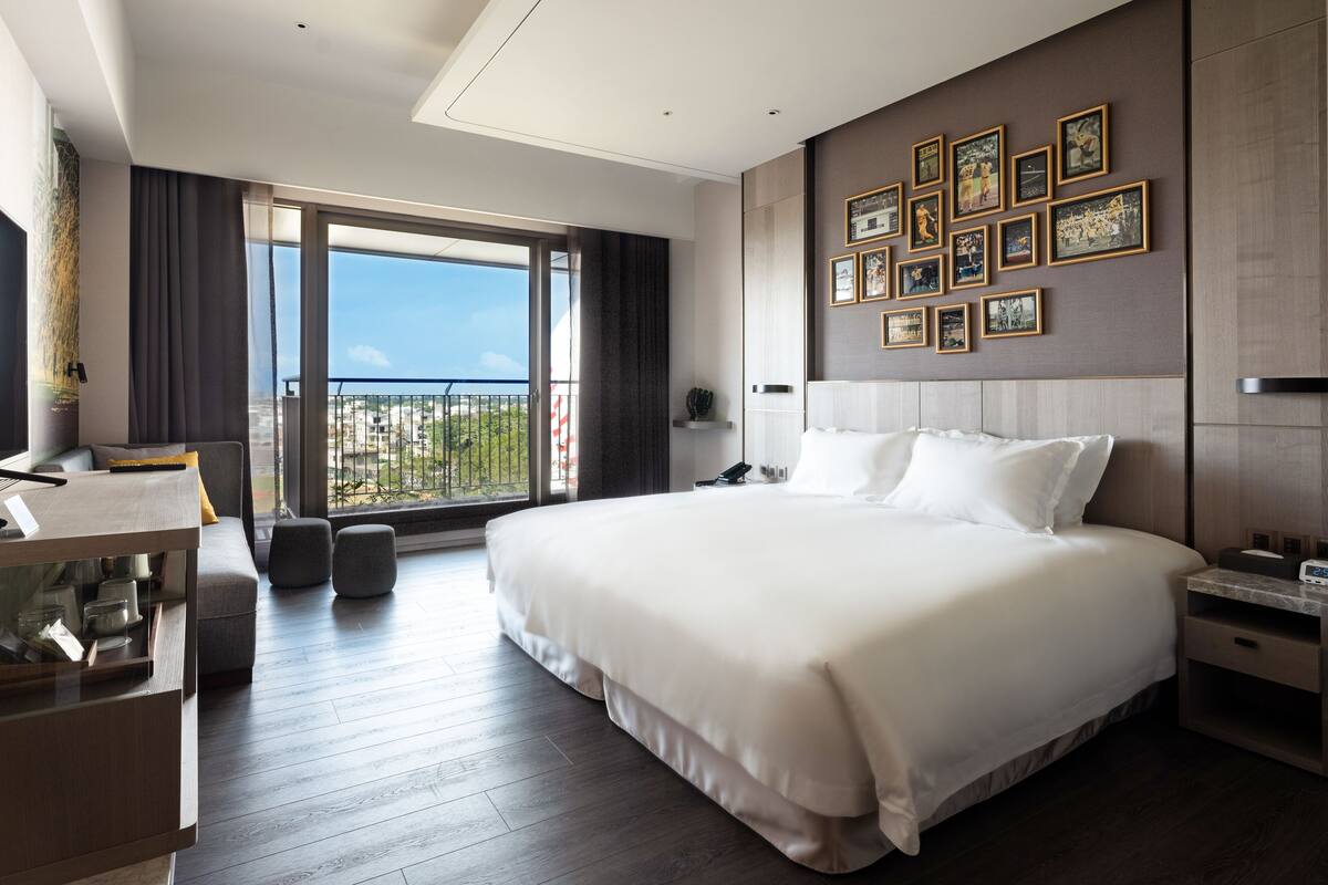 A modern hotel room with a large bed, a desk, and a balcony with a city view.