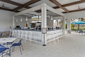 A spacious outdoor bar area with white bar stools and tables, a counter with a drink menu, and a view of the outdoors.