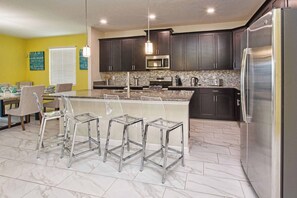 A modern kitchen with dark cabinets, a stainless steel refrigerator, a microwave, and a granite countertop. There are three clear glass stools and a dining table with chairs. - Spacious Storey Lake Home: Walk to Resort, BBQ, Game Room & Free WiFi! (Kissimmee)