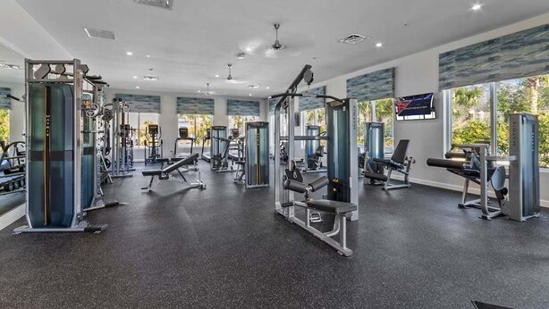 Un gimnasio con diversas máquinas de ejercicio, amplias ventanas e iluminación empotrada en el techo.