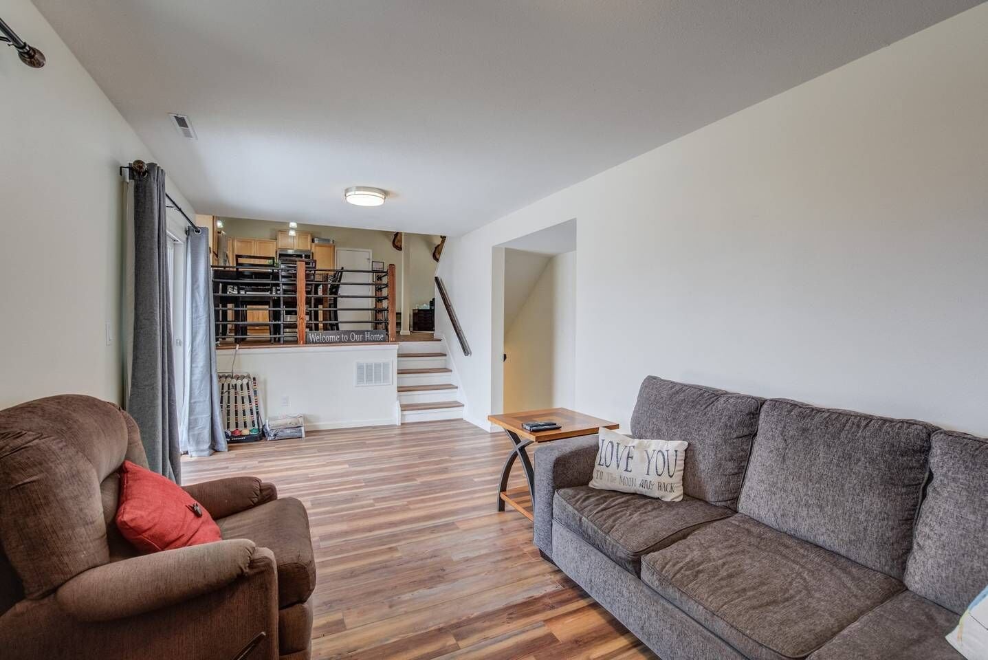 Ein modernes Wohnzimmer mit grauem Sofa, braunem Sessel und Couchtisch aus Holz. Eine Treppe führt zu einer Küchenzeile, an deren Wand ein Schild mit der Aufschrift 'Welcome to Our Home' befestigt ist.