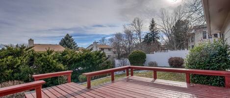 Una terraza de madera con barandas rojas, rodeada de vegetación y edificios residenciales.