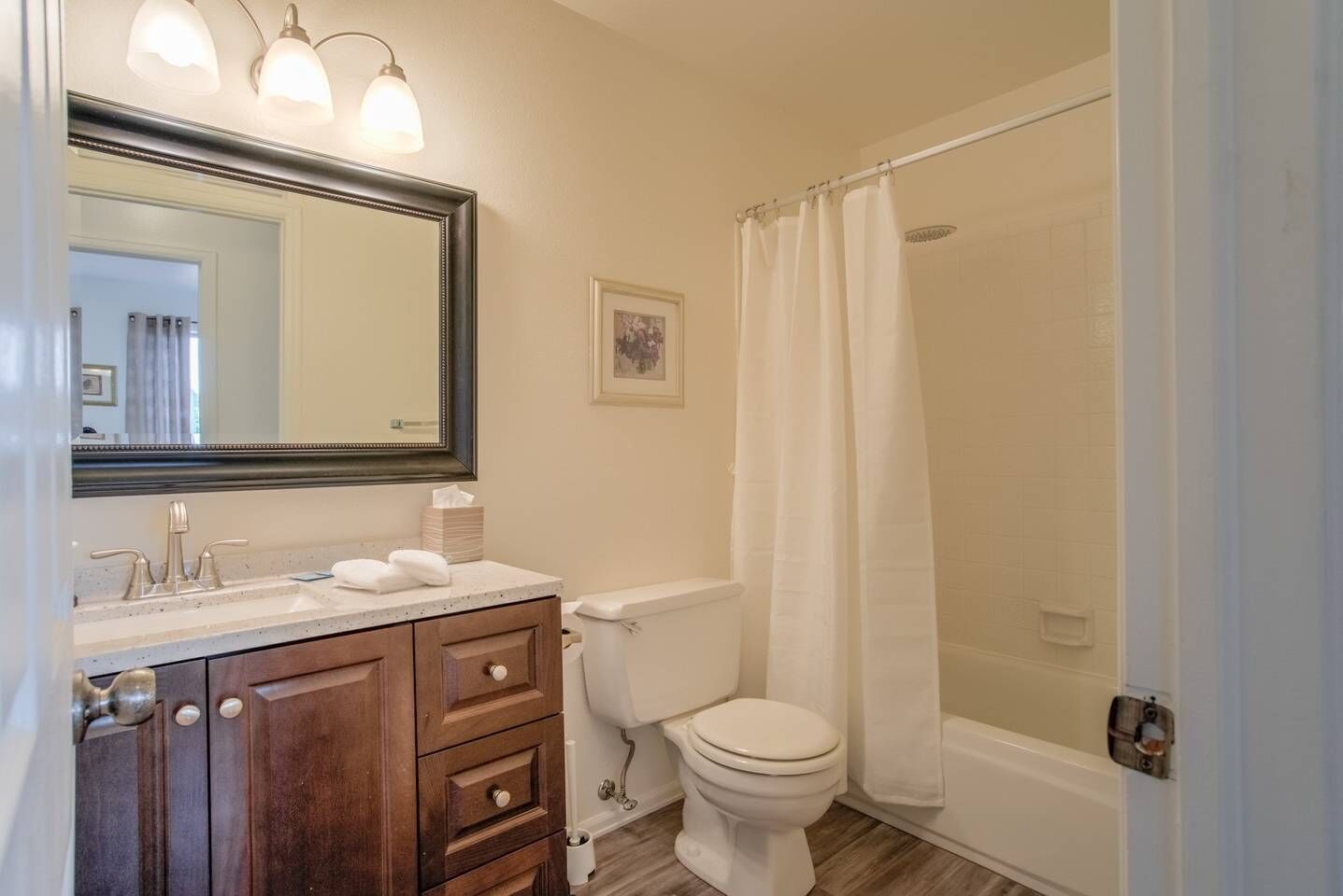 Un baño con tocador de madera, inodoro blanco, ducha con cortina blanca y espejo con luces.