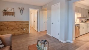 A modern apartment with a wooden accent wall, a dining area with a coffee table, and a kitchen with white cabinets. - Westside Value Parks & Trails Close to it all (Colorado Springs)