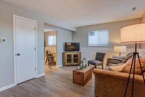 A living room with a sofa, a TV on a wooden cabinet, a dining area with a table and chairs, and a lamp. - Westside Value Parks & Trails Close to it all (Colorado Springs)