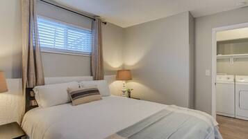 A bedroom with a bed, a nightstand, a lamp, a window with curtains, and a laundry area.