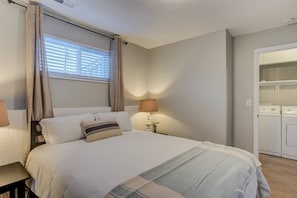 A bedroom with a bed, a nightstand, a lamp, a window with curtains, and a laundry area. - Westside Value Parks & Trails Close to it all (Colorado Springs)
