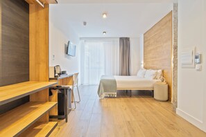 A modern hotel room with a wooden floor, a bed with a white headboard, a desk with a chair, a TV mounted on the wall, and a large window with curtains. - Colectia Hotel Ondarreta (San Sebastián)