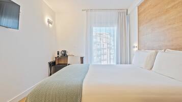 A hotel room with a large bed, a desk with a chair, a coffee maker, and a view of buildings through sheer curtains.