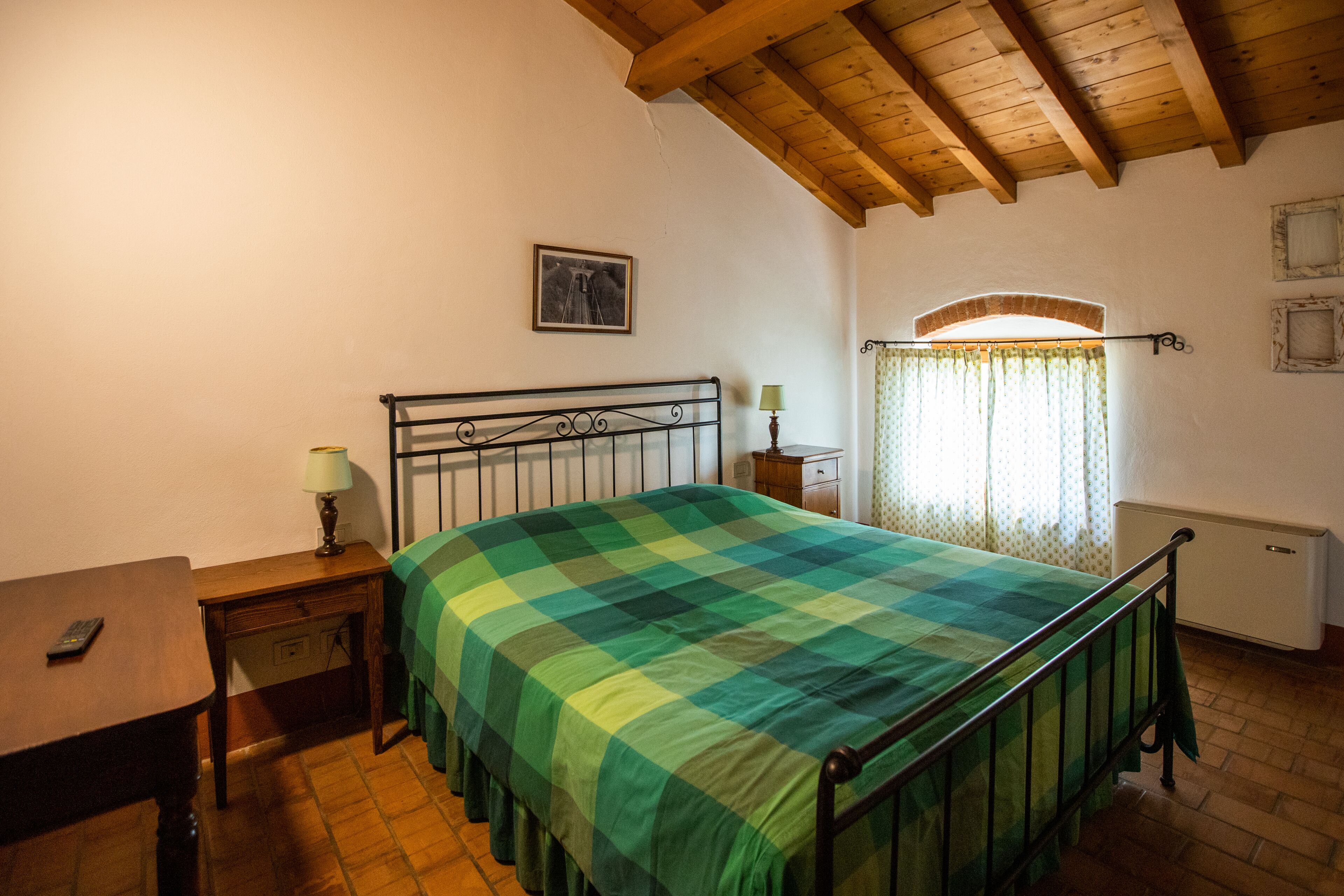Una camera da letto con un letto, un comodino, una lampada, una finestra con tende e un quadro appeso al muro.