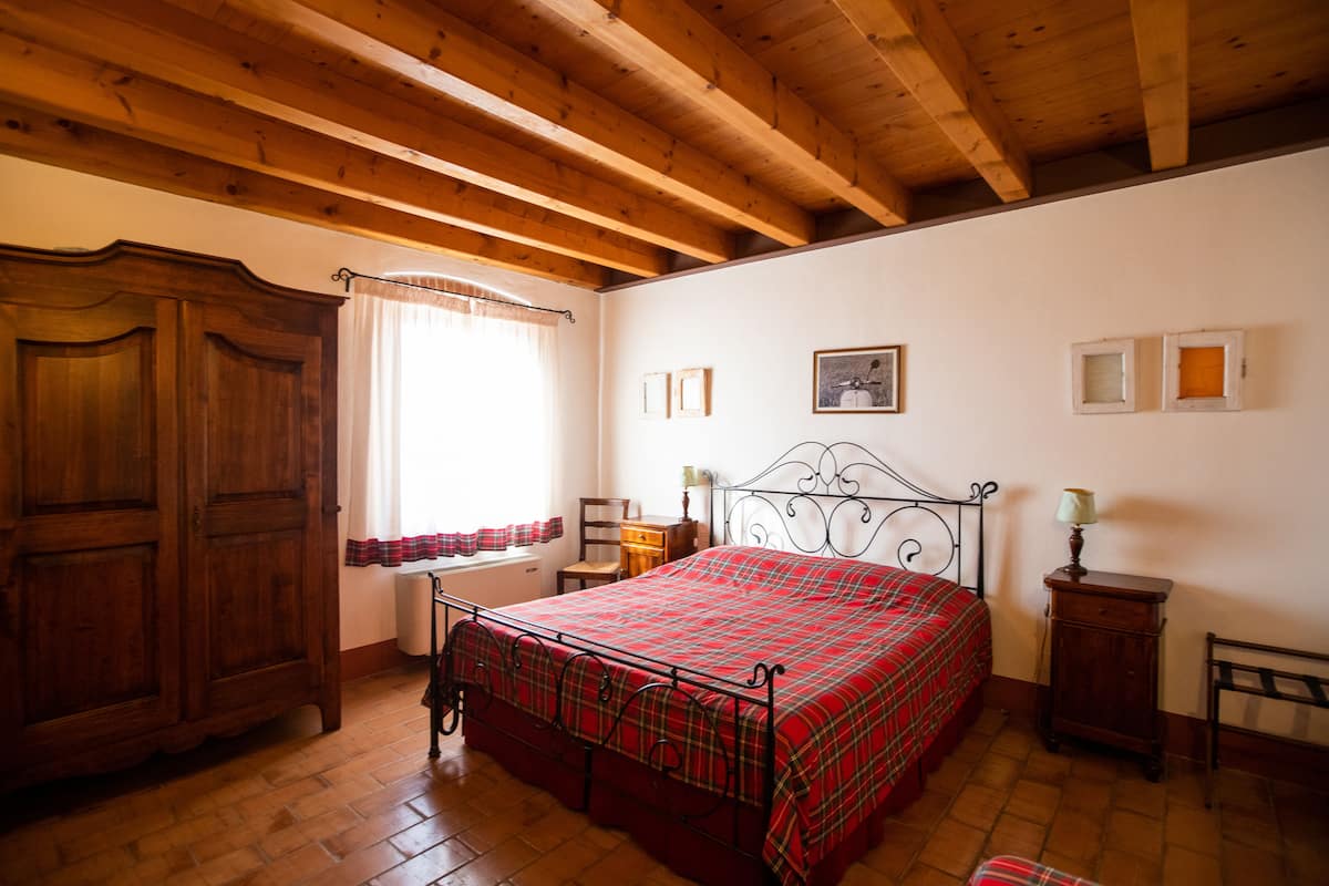 A bedroom with a wooden ceiling, a bed with a red plaid bedspread, a wooden wardrobe, a window with curtains, and a small bedside table with a lamp.