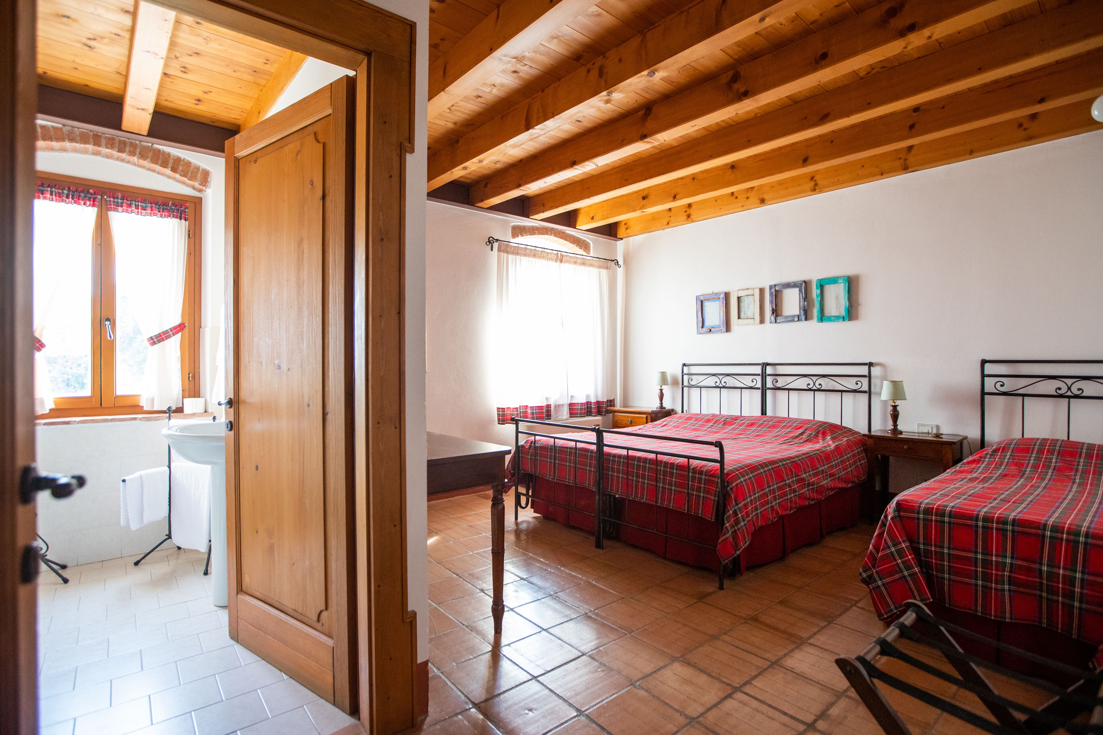 A room with two beds, a wooden ceiling, a window with curtains, and a bathroom with a sink and towel rack.