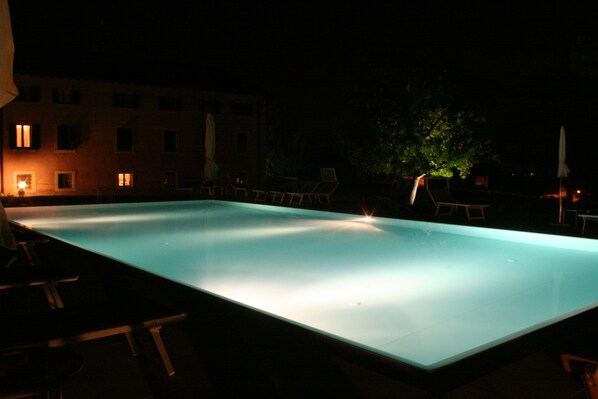 A rectangular swimming pool at night with surrounding lounge chairs and a building in the background. - Agriturismo Ca' Vecia (Sona)