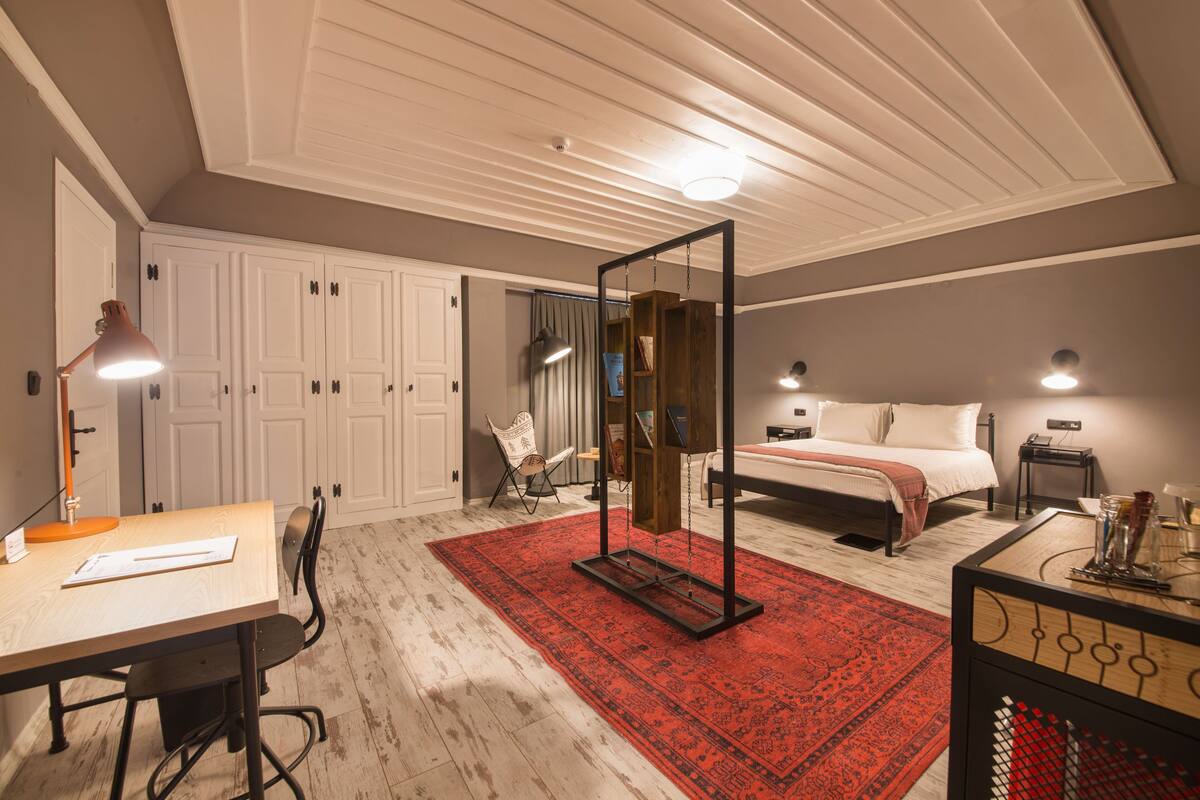 Un dormitorio con cama, escritorio con lámpara, silla, armario y alfombra roja estampada.