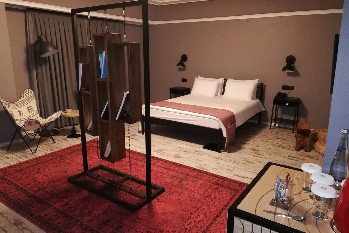 A hotel room with a bed, a red rug, a hanging chair, a small table with glasses and a bottle, and a wooden shelf with books.