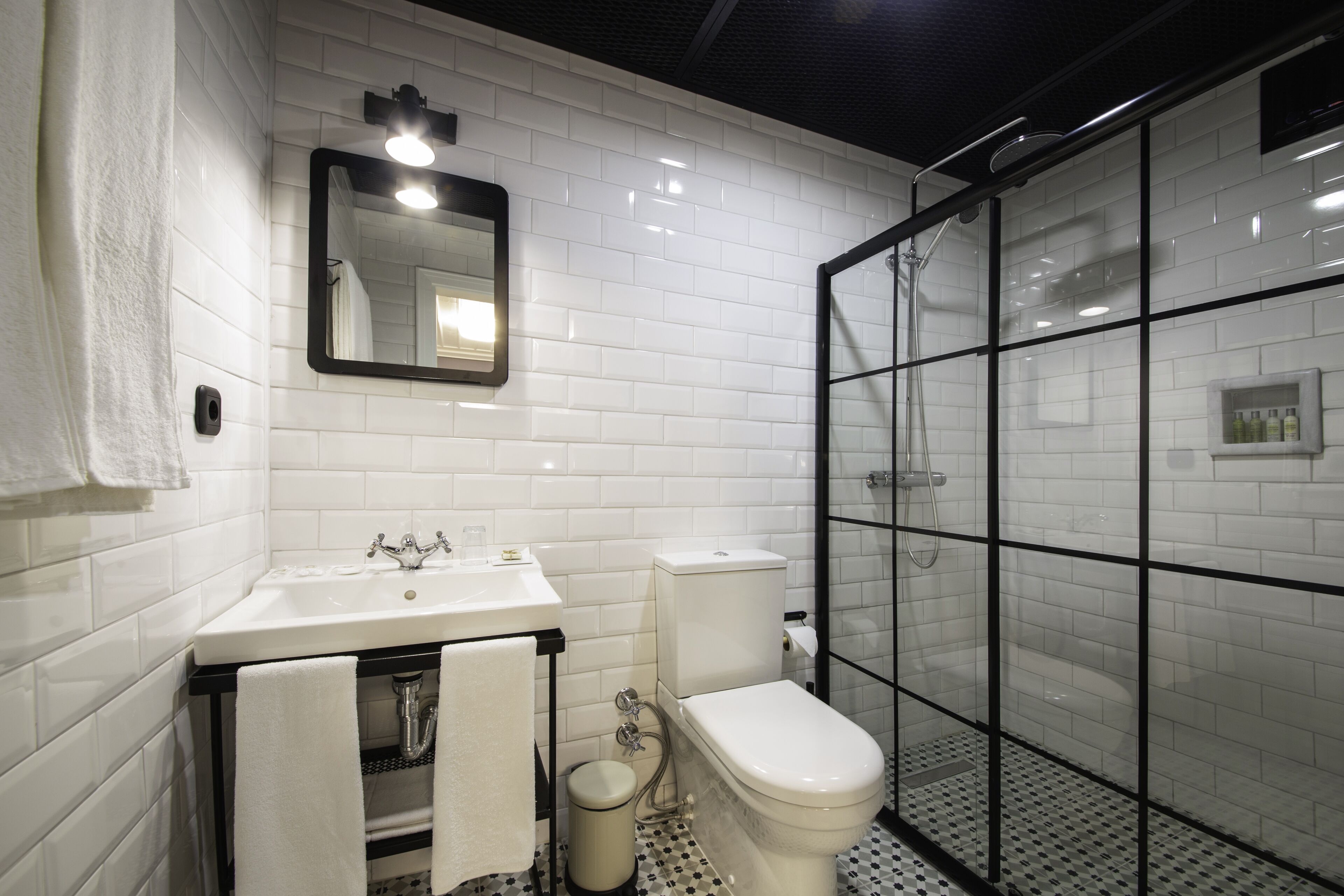 Un baño moderno con azulejos blancos tipo metro, una ducha con mampara de vidrio, un lavamanos blanco y un inodoro.