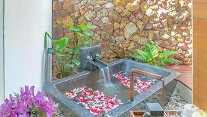 A spa area with a stone wall, a hot tub with rose petals, and a wooden tray with toiletries.