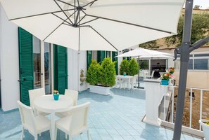 A covered outdoor patio with white wicker furniture, green shutters, and potted plants. - Casa MareLuna (Vico Equense)