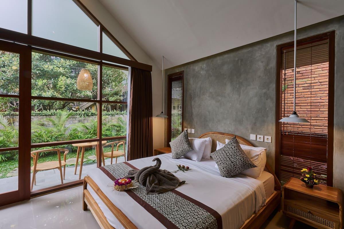 A modern bedroom with a large window, a bed with patterned bedding, and a small table with a lamp.