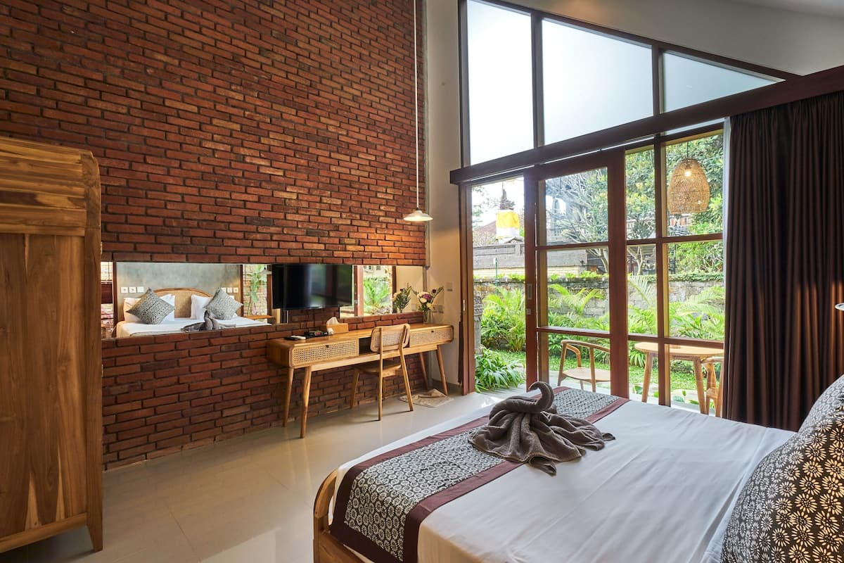 A bedroom with a brick wall, a large window, a bed, a desk, and a TV.