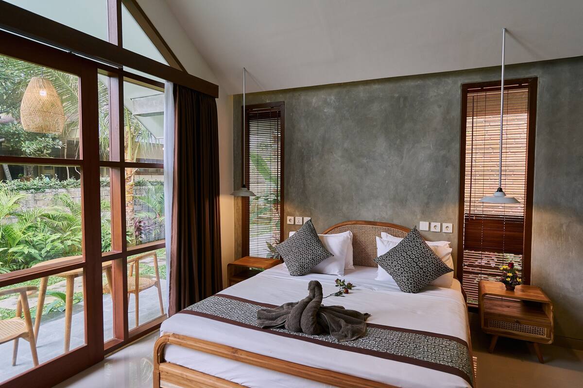 A bedroom with a large window, a bed with pillows, a bedside table with a lamp, and a view of greenery outside.