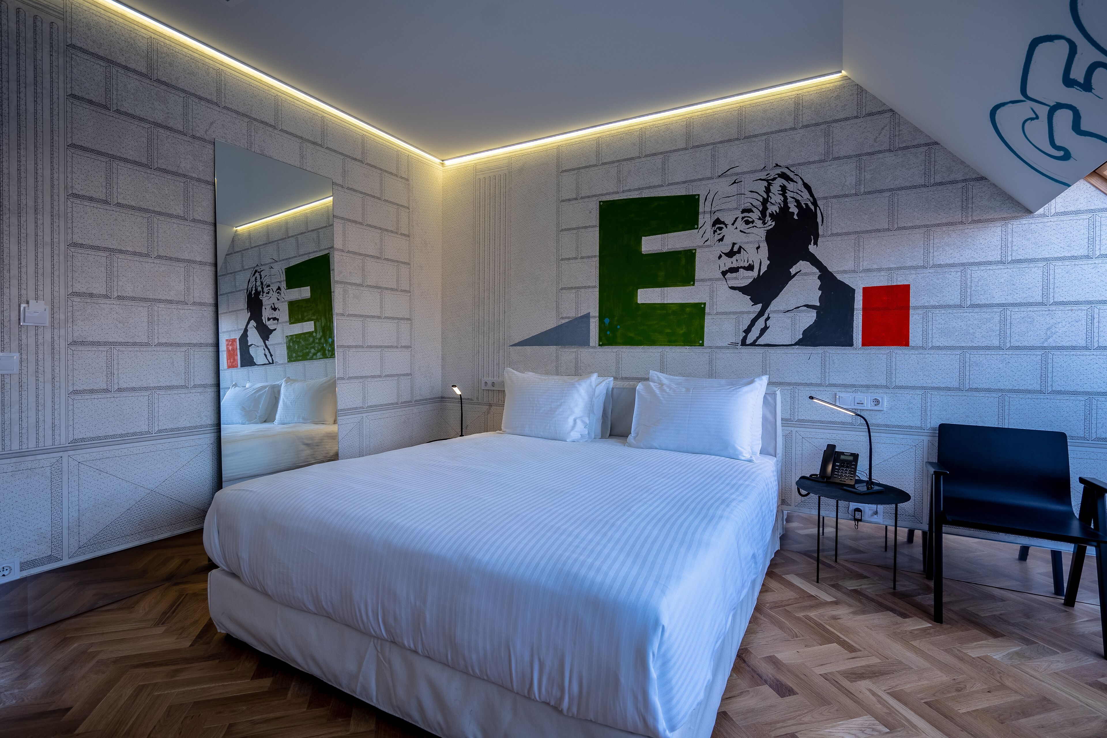 A modern hotel room with a large bed, a chair, a desk with a lamp, and a mirror on the wall.