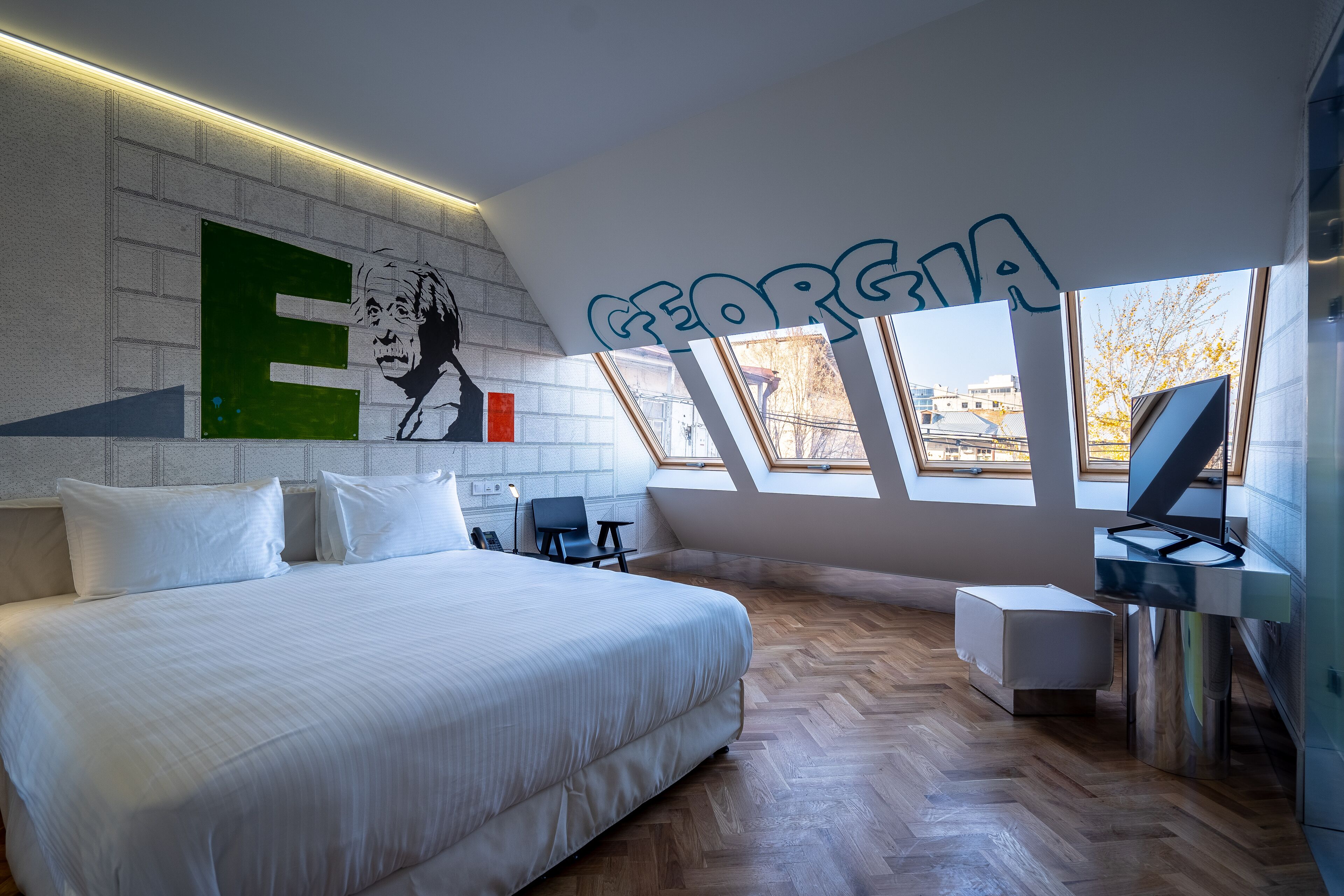 Ein modernes Schlafzimmer mit einem großen Bett, einem Schreibtisch und einem Computer. An der Wand befindet sich ein großes 'E' und ein Fenster mit Blick auf Gebäude.