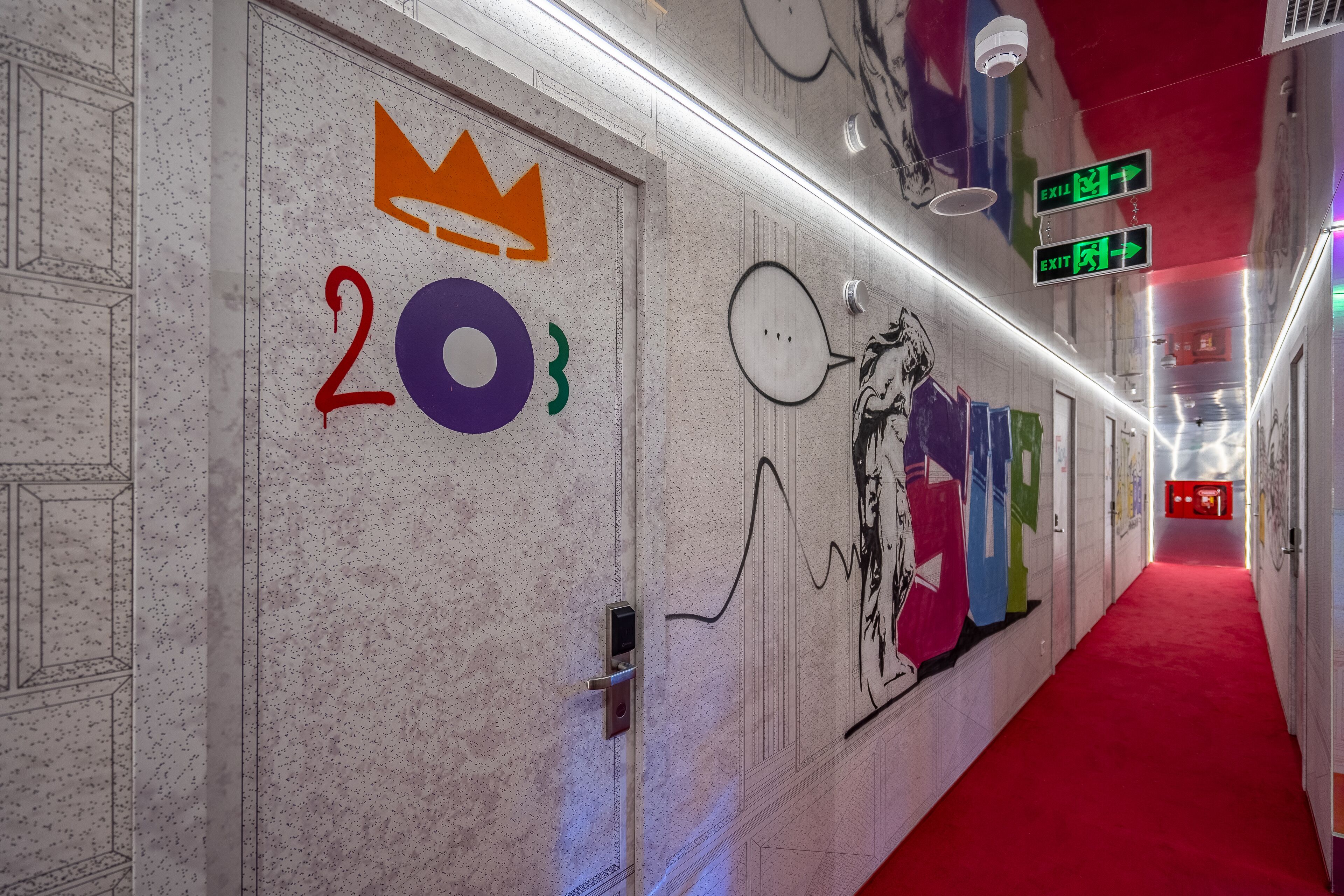 Un pasillo con alfombra roja, una puerta con el número 203 y un símbolo de corona, y una pared con grafitis coloridos y un bocadillo de diálogo.