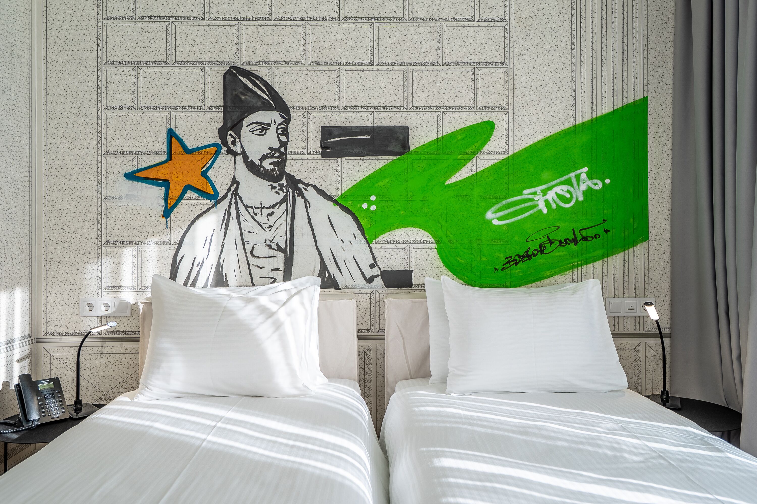 A hotel room with two beds, a nightstand with a phone, and a wall with a mural and a green sign.