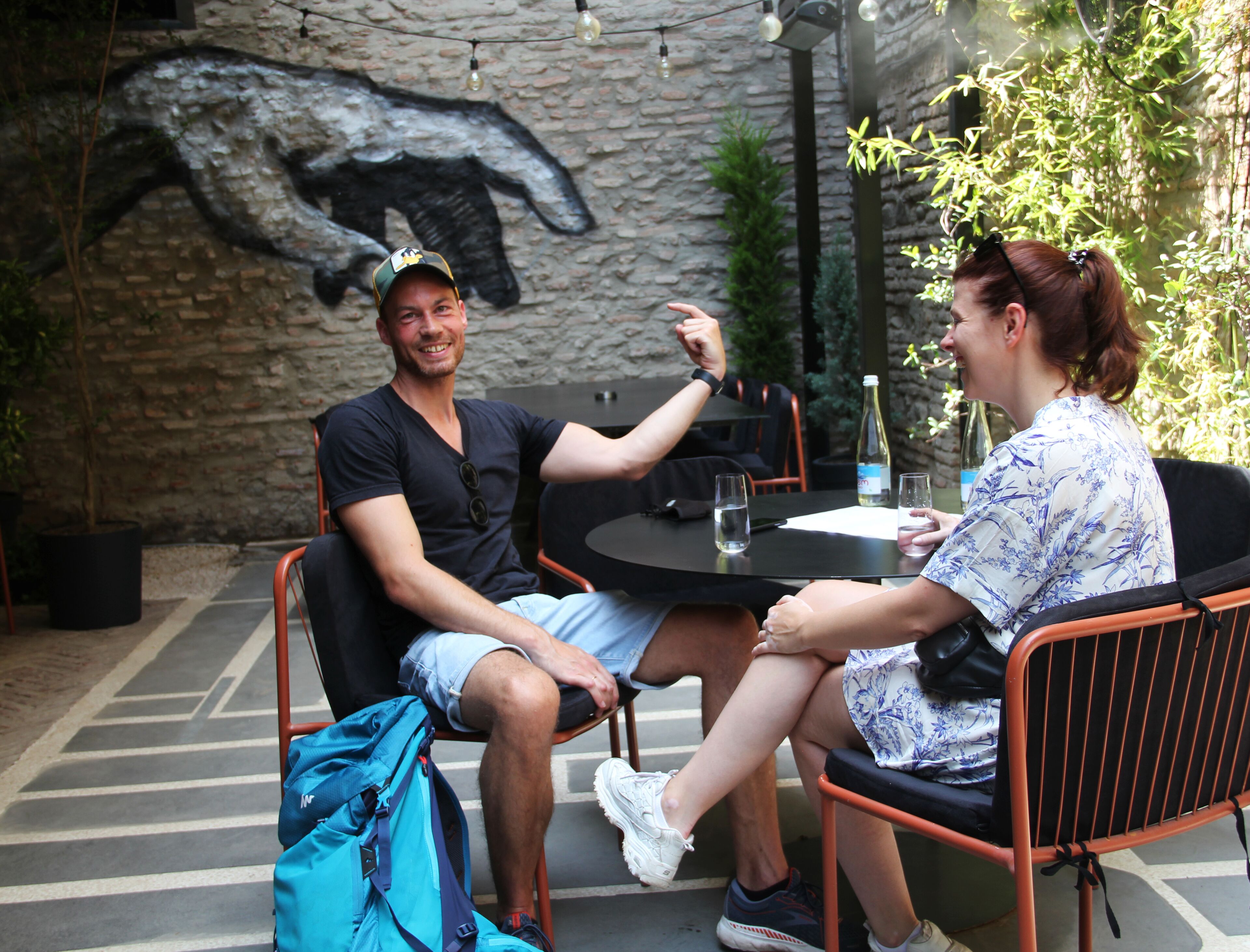 Two people seated at an outdoor table with a stone wall mural in the background.