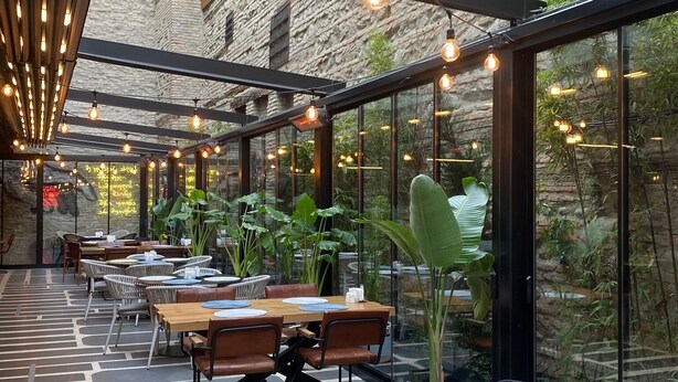 Un comedor interior con mesas y sillas, ventanas amplias y luces colgantes.