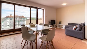 A modern living room with a dining area, large windows, a grey sofa, and a flat-screen TV. - Victus Apartamenty - Cadena 1 (Sopot)