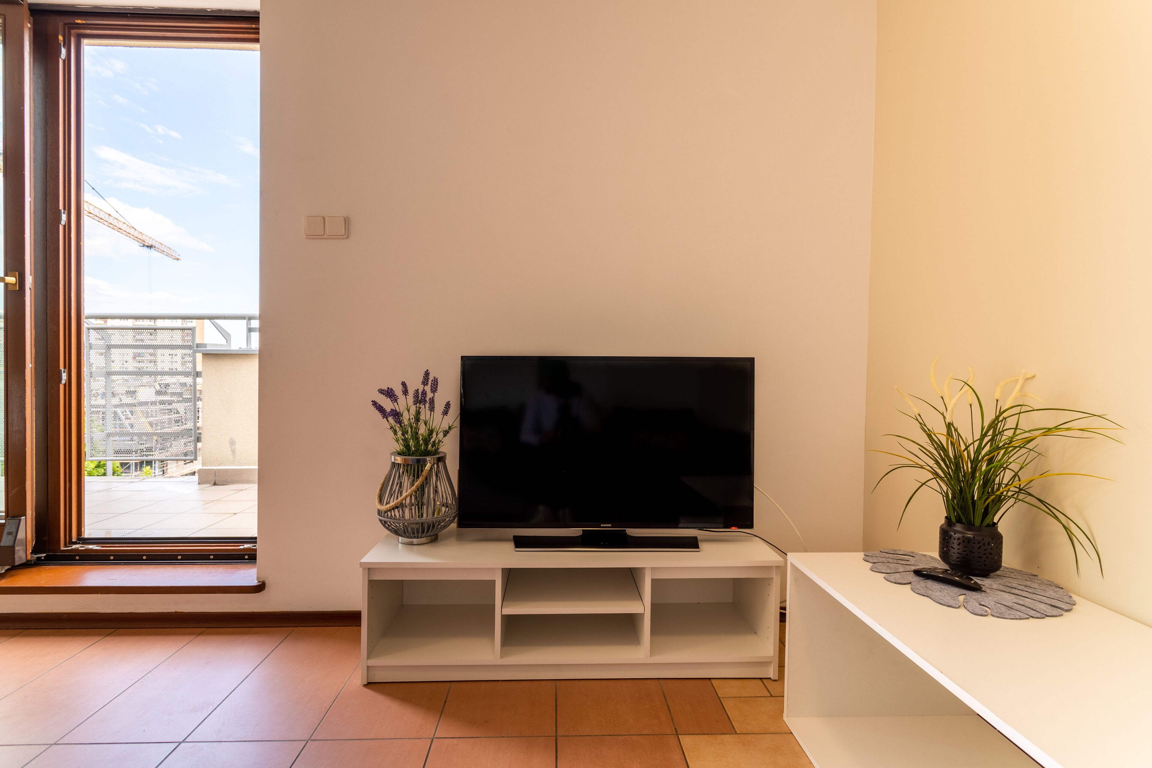 A living room with a flat-screen TV on a white entertainment unit, a potted plant, and a view of a balcony with a building in the background.