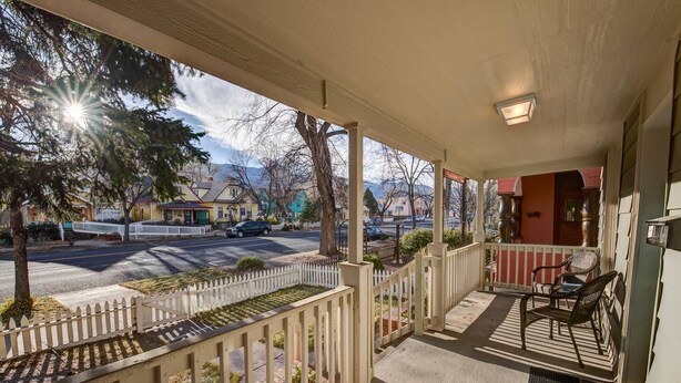 A porch with a white picket fence, outdoor seating, and a view of a street with cars and houses.
