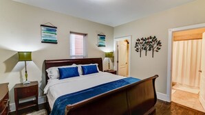 A bedroom with a wooden bed, two bedside tables with lamps, a window with blinds, and a door leading to another room.