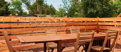 Une terrasse extérieure en bois, dotée d’une table et de chaises intégrées, entourée d’une clôture en bois et illuminée de guirlandes lumineuses.