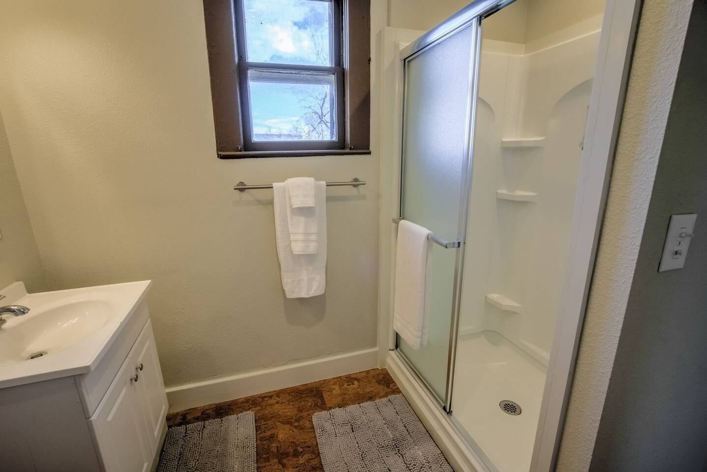 Une salle de bain avec un lavabo blanc, une cabine de douche en verre et une fenêtre au-dessus du lavabo.