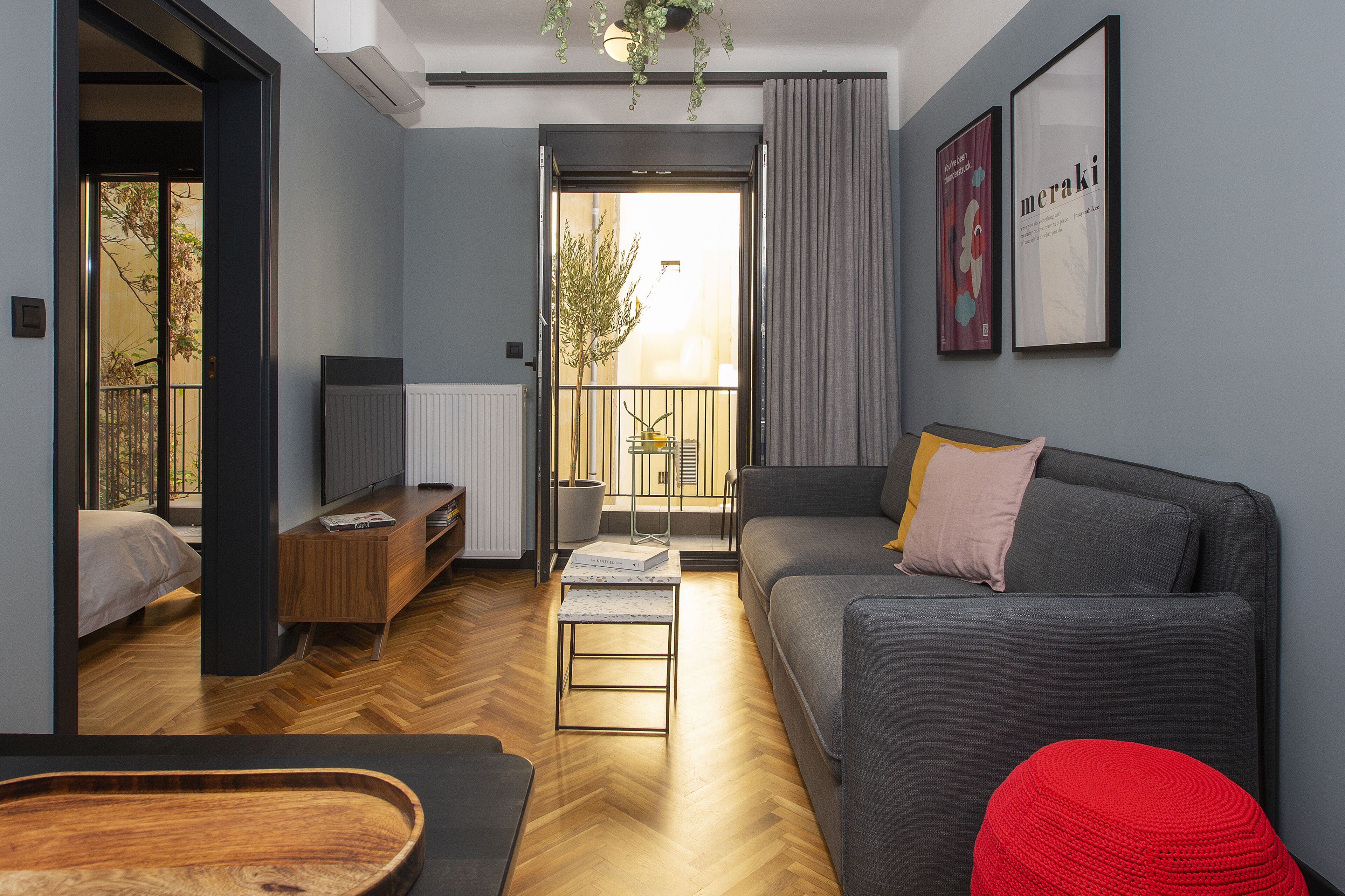 A modern living room with a grey sofa, a red ottoman, a wooden coffee table, and a small white table. There is a balcony with a view of trees and a building.