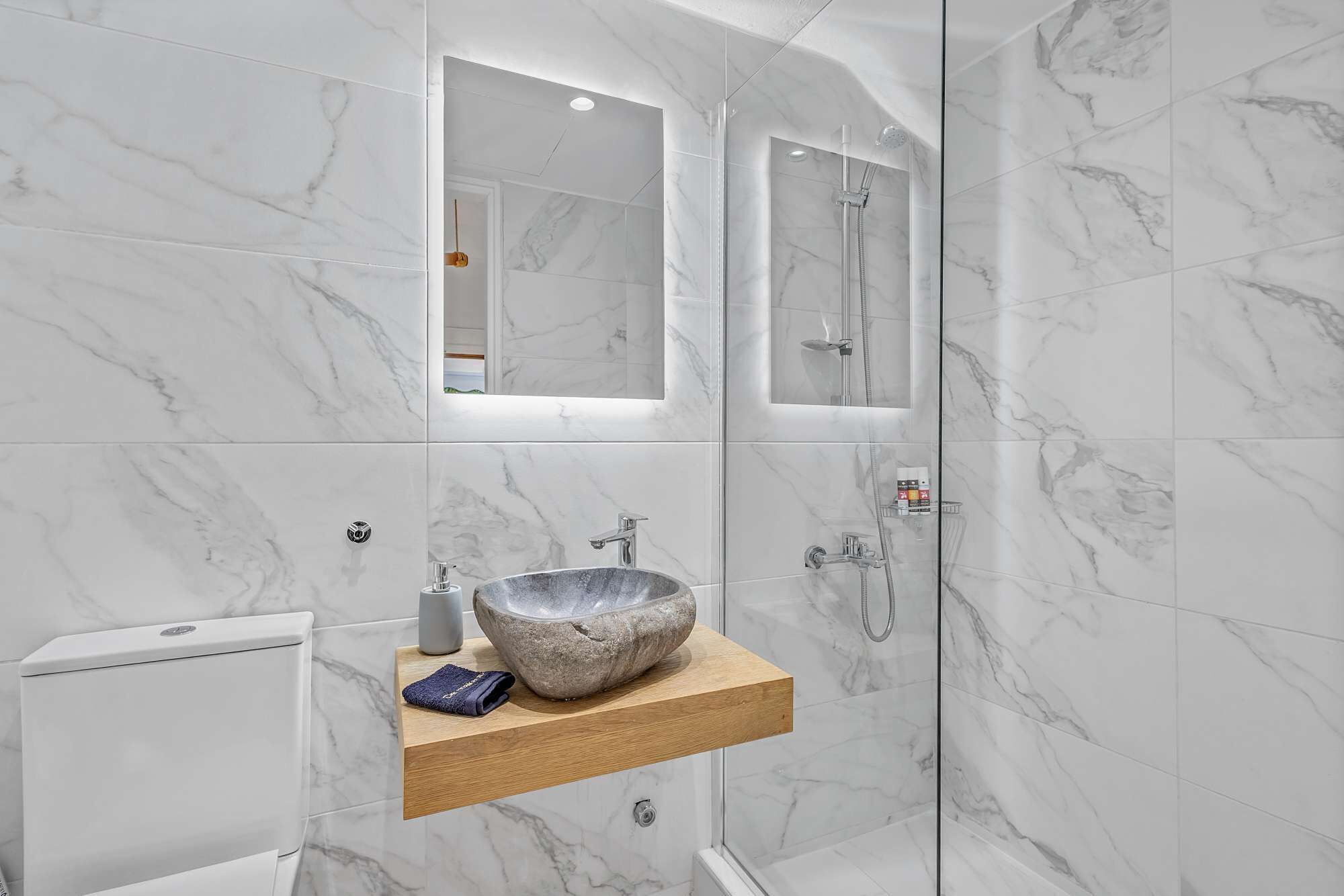 Een moderne badkamer met een betegelde wand in marmerlook, een houten wastafelmeubel, een stenen wastafel en een douchecabine met glazen wanden.