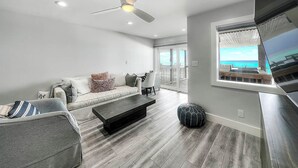 A modern living room with a sofa, coffee table, and ottoman. There is a ceiling fan, a window with a view of the ocean, and a balcony with a railing. - Into the Blue - Oceanfront Condo with Beach Access & Gulf Views (Miramar Beach)