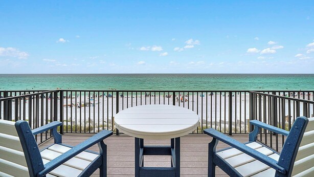 A deck with a table and two chairs, overlooking a beach with people and a clear sky. - Into the Blue - Oceanfront Condo with Beach Access & Gulf Views (Miramar Beach)