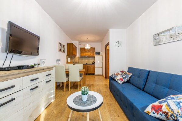 A modern living room with a blue sofa, a flat-screen TV, a wooden dining table, and a kitchen in the background.
