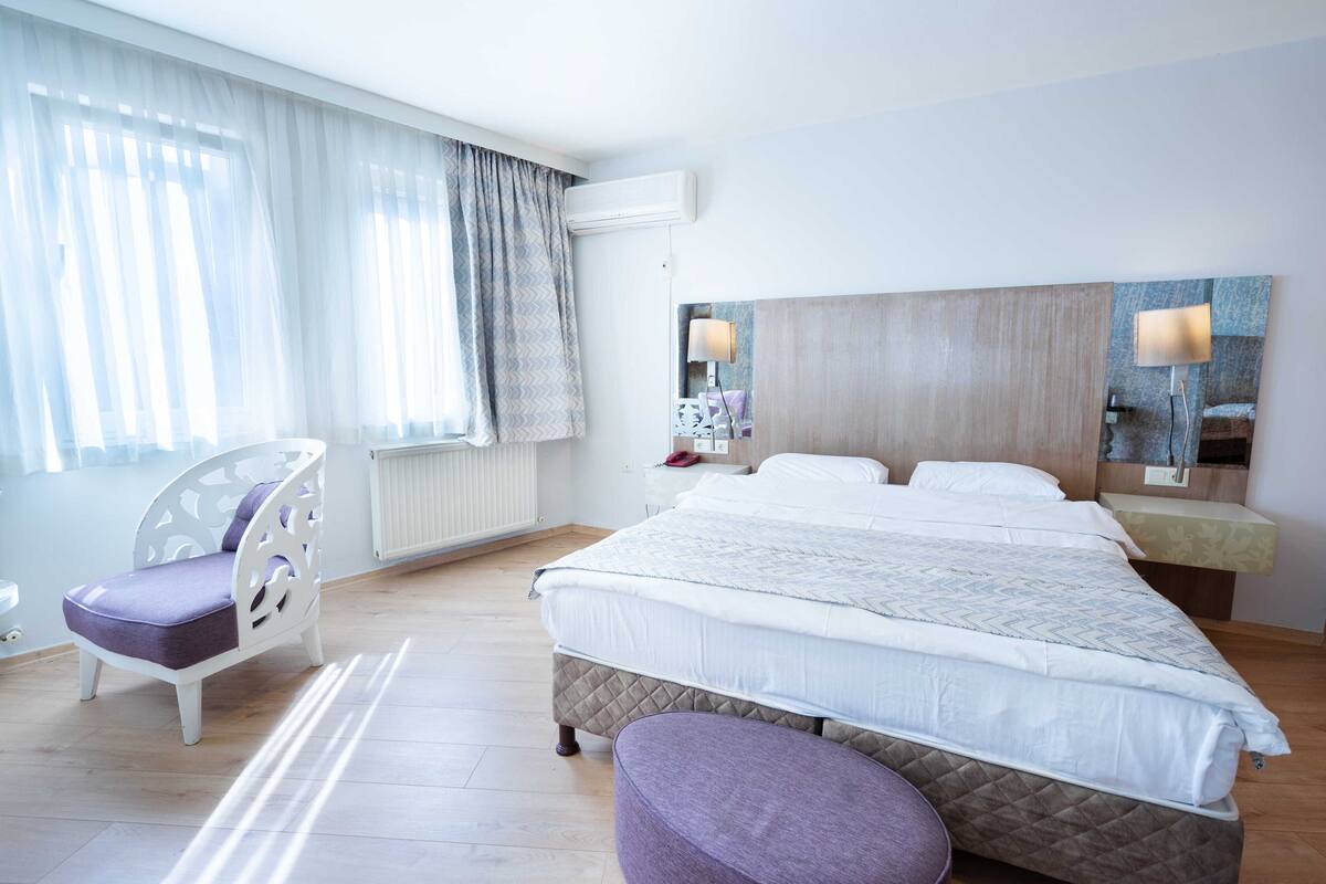 A hotel room with a large bed, a chair, a bedside table, and a wall-mounted air conditioner.