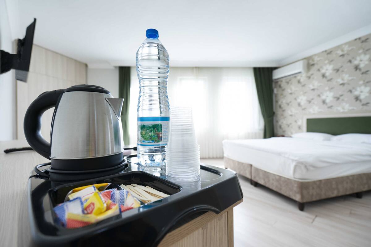 A hotel room with a kitchenette area, including a kettle, a water bottle, and stacked plastic cups.