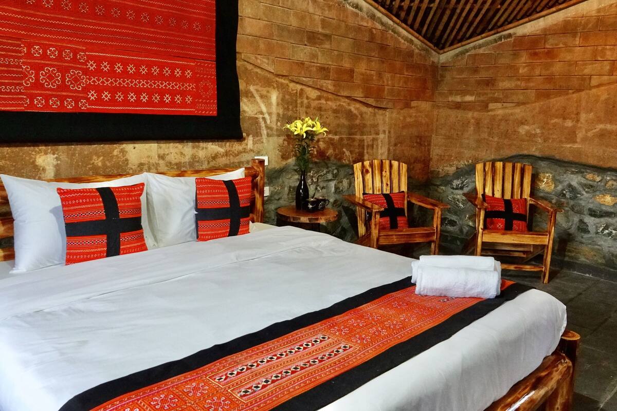 A bed with white and red bedding, two wooden chairs with red cushions, a small table with a vase of flowers, and a stone wall.
