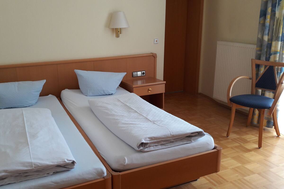 Une chambre d’hôtel avec deux lits simples, chacun muni d’un oreiller et d’une table de chevet. On y trouve également une chaise en bois et un bureau avec une lampe.