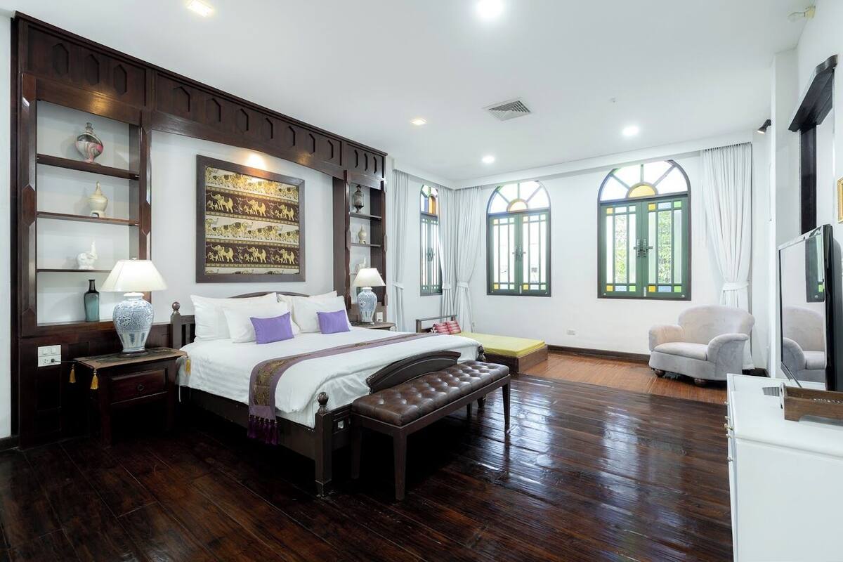 Un dormitorio amplio con una cama grande, un cabecero de madera, una mesita de noche, una lámpara, un banco, una silla, un televisor y una ventana con cortinas.