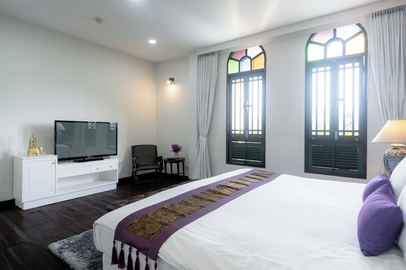 Quarto de hotel com cama, televisão, cadeira, mesinha e janela com cortinas.