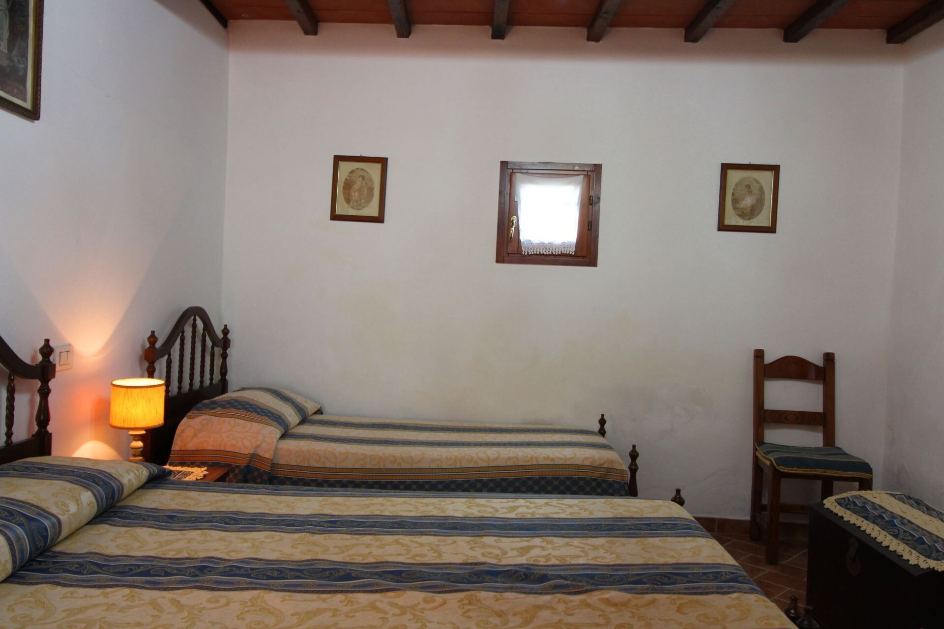 A bedroom with two beds, a wooden headboard, a nightstand with a lamp, a wooden chair, and a small window.