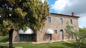 Un edificio de piedra con un gran árbol en el frente, un pequeño jardín y un cielo despejado.